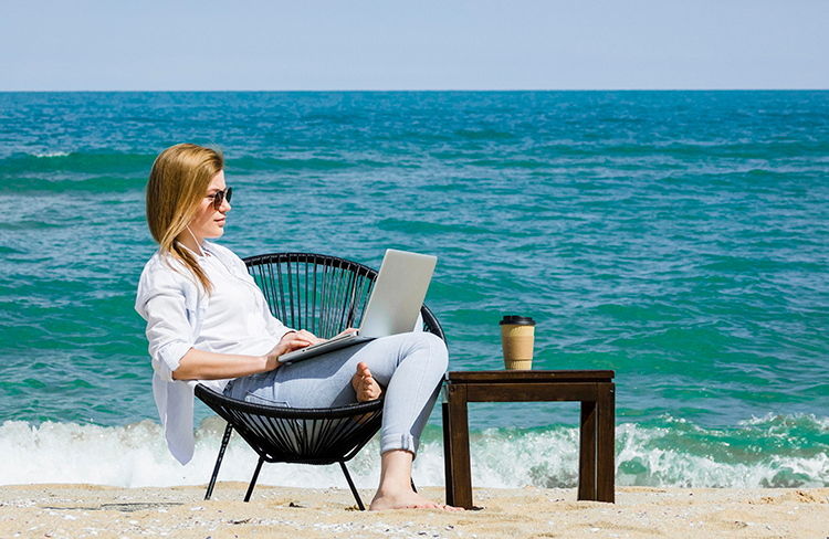 Image of a woman using a laptop on a beach to represent the work anywhere benefit often cited as a reason to move to AVD